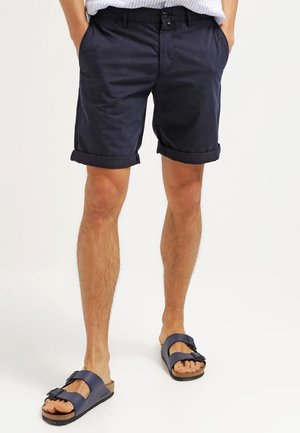 Niebieskie bawełniane szorty z podwiniętymi nogawkami, z zapięciem na guzik z przodu i bocznymi kieszeniami, w połączeniu z ciemnymi sandałami.