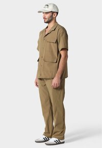 Camisa de manga corta marrón claro con dos bolsillos en el pecho, combinada con pantalones a juego. Lleva una gorra blanca con un logo y rayas negras en los zapatos.