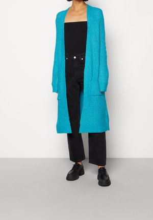 Persona che indossa un lungo cardigan di maglia turchese sopra una canotta nera senza spalline, jeans neri a vita alta e scarpe nere con plateau e lacci.