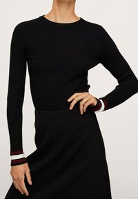 Haut noir à manches longues en côtes, avec un col rond ; présente des accents rayés rouges et blancs au niveau des poignets. Texture lisse et design ajusté.