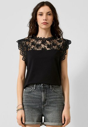 Femme portant un haut noir sans manches avec un détail en dentelle florale aux épaules et au col, associé à un short en jean gris retroussé.