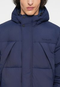 Abrigo acolchado de un azul marino oscuro con cierre de cremallera y botones en la parte frontal, que presenta un cuello alto, capucha ajustable y costuras visibles para aislamiento.
