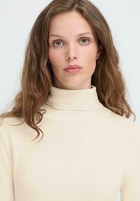 Beżowy sweter z golfem o miękkiej fakturze, dopasowanym kroju i długich rękawach, noszony przez osobę z falującymi brązowymi włosami i naturalnym makijażem.