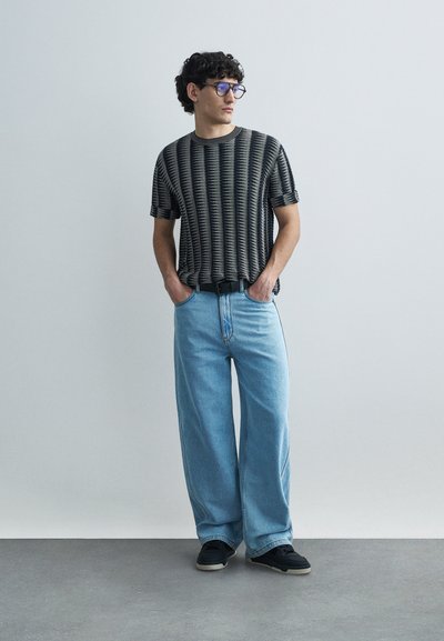 T-shirt à manches courtes à rayures noires et crème, associé à un jean large bleu clair et des baskets noires. Accessoirisé avec des lunettes bleues.