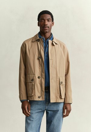 Man wearing tan buttoned jacket over blue polo shirt and blue jeans standing against plain light background.