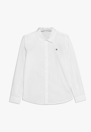 Camisa blanca de manga larga y botón en algodón, con un cuello clásico y un pequeño logo negro de CK en el lado izquierdo del pecho. Textura suave.