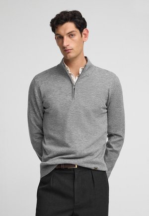 Grijze zip-neksweater gemaakt van zacht materiaal, met lange mouwen en een relaxte pasvorm, gecombineerd met donkere broeken en een verborgen knoop-down kraag.