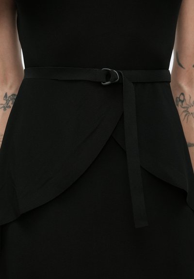 Czarna kopertowa sukienka z cienkim paskiem zawiązanym na supeł w talii, odsłaniająca tatuaże na przedramionach po obu stronach.