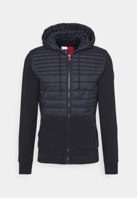 Felpa con zip color navy, con corpo imbottito; presenta un cappuccio, maniche lunghe, polsini a costine e un logo accentuato sull'orlo. Materiali lisci e testurizzati.