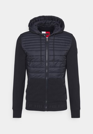 Navy zip-up hoodie with padded body; features a hood, long sleeves, ribbed cuffs, and a logo accent on the hem. Smooth and textured materials.