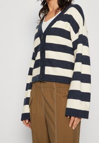 Marinblå och krämrandig cardigan med V-ringning, fem knappar, lösa ärmar och i en blandning av ullmaterial. Matchas med bruna byxor.