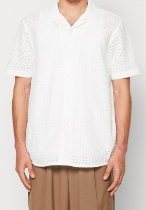 Mężczyzna ubrany w białą, krótką koszulę zapinaną na guziki z ażurowego materiału oraz beżowe plisowane spodnie, stojący na tle jednolitego tła.