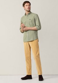 Camisa de botón en tela verde claro, con un bolsillo en el pecho; combinada con pantalones amarillos y zapatos marrón oscuro. Corte sencillo y moderno.