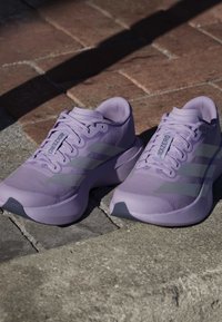 Para lawendowych butów do biegania Adidas Adizero umieszczona na brukowanym zewnętrznym terenie ze światłem słonecznym i cieniami.