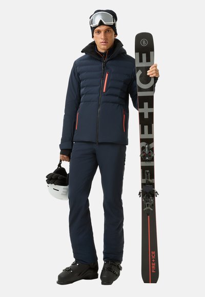 Navy Skijacke im Pufferdesign mit orangefarbenen Akzenten, kombiniert mit passenden Hosen. Hält schwarze Skier mit Logo und weißen Helm.