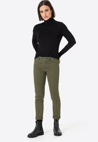 Zwarte mock turtleneck top gecombineerd met olijfgroene slim-fit broek. Broek heeft vijf zakken en een knopenfly. Gecombineerd met zwarte enkelboots.