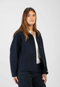 Giacca in lana di un blu scuro con una vestibilità rilassata, caratterizzata da una chiusura con bottone, due tasche e un design leggermente accorciato. Abbinata a una camicia bianca.