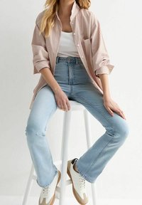 Žena v světle modrých rozšířených džínách, bílých teniskách, bílé tílku a přes oversized růžovém pruhovaném triku sedí na bílém stoličce.