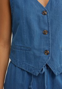 Modra denim križna jakna z globokim V-izrezom, štirimi rjavimi gumbi in stranskimi žepi. Tkanina ima mehak otip in rahlo strukturirano obliko.