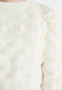 Pull blanc tricoté présentant un motif zigzag texturé, avec un matériau doux et moelleux et un col côtelé. Manches détaillées avec une légère ampleur.