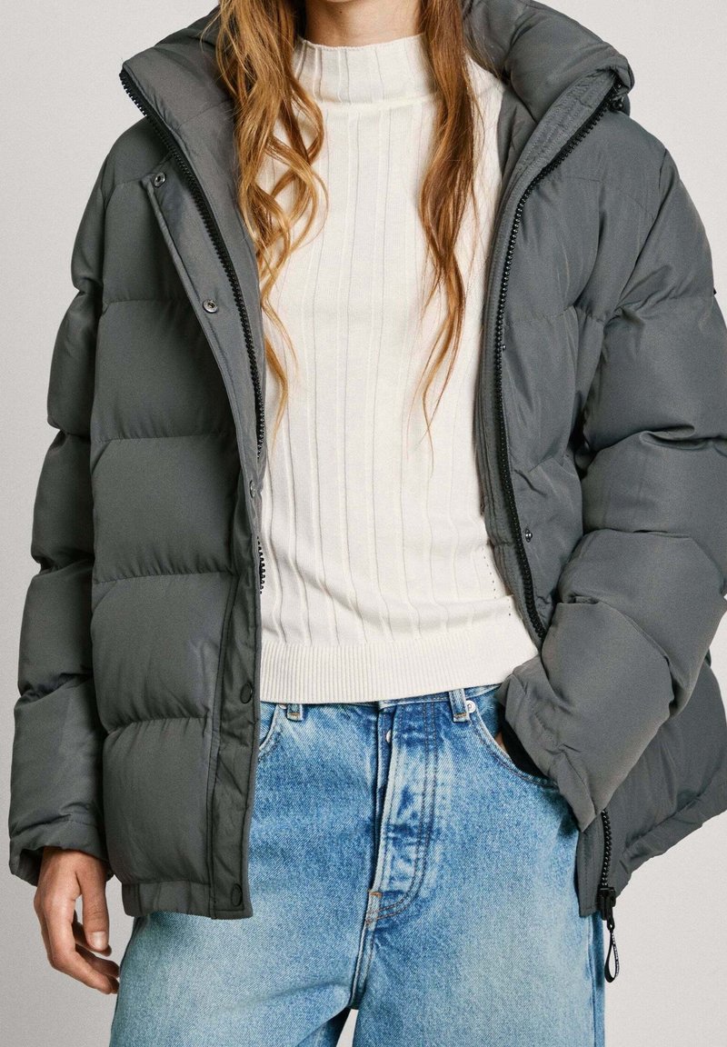 Giacca imbottita grigia con colletto alto, dotata di cerniera completa e bottoni automatici, indossata sopra un maglione bianco a coste e jeans azzurri.