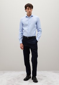 Giovane uomo in piedi con una camicia azzurro chiaro, pantaloni blu scuro, cintura nera e scarpe eleganti nere su un tappeto chiaro.