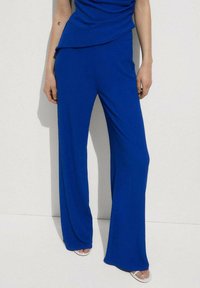 Blue wide-leg pants made of soft fabric, featuring a draped waistband and smooth texture. Worn with a matching top and minimal footwear.