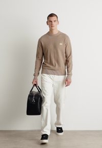Maglione lavorato a maglia beige con scollo rotondo, pantaloni dritti bianchi e sneakers nere; in mano una borsa in pelle nera.