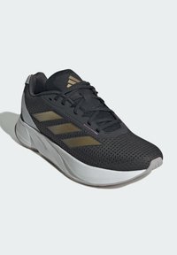 Černé sportovní boty s prodyšným síťovaným svrškem, stříbrnými akcenty a třemi zlatými pruhy. Podrážka s polstrovaným designem a texturovanými vzory.