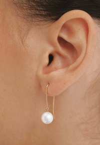 Pendientes de metal color dorado con un diseño de gancho curvado, que presentan una sola perla blanca al final, suspendida debajo de la oreja.