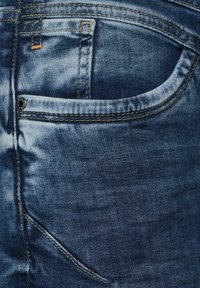 Džínové kalhoty v tmavě modré barvě s vybledlým efektem. Mají přední kapsu s kovovým knoflíkem a kontrastním žlutým prošíváním podél švů.