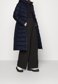 Manteau matelassé long bleu marine avec capuche, présentant un design matelassé, des finitions contrastées, des poches latérales et une coupe décontractée, associé à un pantalon large noir.