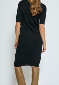 Svart midiklänning med korta ärmar och lös passform. Slät tyg med en enkel struktur. Modellen har på sig knähöga beige stövlar.