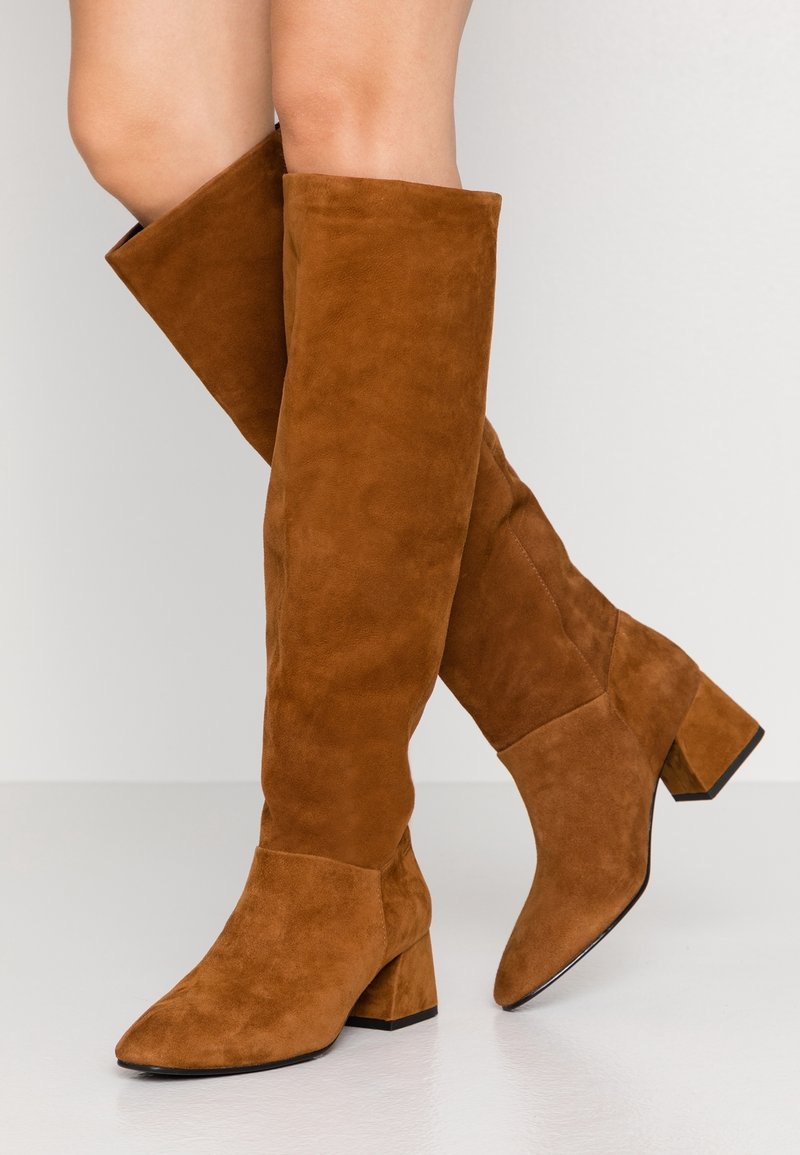 Mujer con botas de ante marrón hasta la rodilla y tacones cuadrados, de pie con las piernas cruzadas sobre un suelo blanco contra una pared clara.