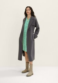 Grauer Wollmantel über grünem Blumenkleid mit langen Ärmeln, doppelter Knopfleiste und beigen Knöchelstiefeln. Schlichtes, geradliniges Design.