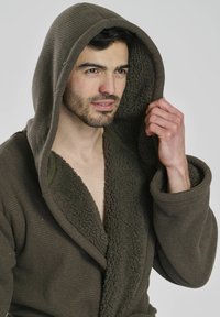 Braune Kapuzenrobe mit Waffelstruktur und weicher Sherpa-Fütterung. Verfügt über eine große Kapuze und übergroße Ärmel. Offenes Frontdesign.