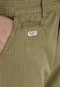 Close-up of beige patterned pants with a small white label reading "BLEND DENIM" near the front pocket with a hand inside.