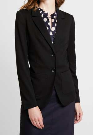 Kvinde iført sort skræddersyet blazer over en navy nederdel og polkaprikket bluse, stående mod en ensartet lys baggrund.