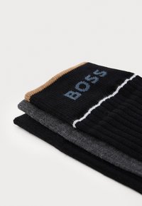Tre par av ribbad strumpor i svart, grått och brunt, med "BOSS"-logotyp på den övre svarta strumpan. Tillverkade av stickat tyg med kontrasterande färger.