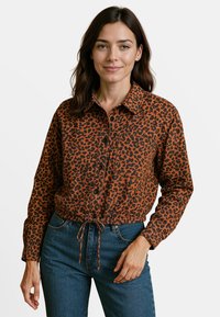 Tijgermotief shirt in oranje en zwarte stof, knoopsluiting aan de voorkant, kraag, borstzak en gathered zoom met een trekkoord. Blauwe spijkerbroek.