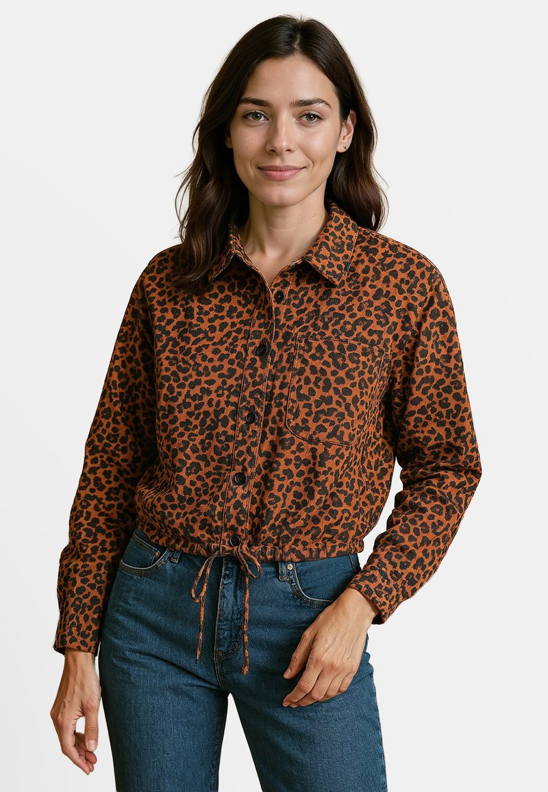 Tijgermotief shirt in oranje en zwarte stof, knoopsluiting aan de voorkant, kraag, borstzak en gathered zoom met een trekkoord. Blauwe spijkerbroek.