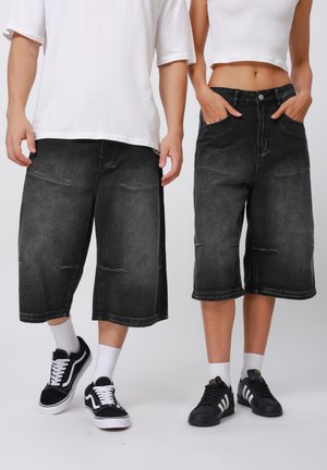 Pantalones cortos de mezclilla negros con un corte holgado, hasta la rodilla, con bolsillos frontales y dobladillos deshilachados. Llevados con zapatillas negras y calcetines blancos.