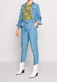 Dżinsowa kurtka, kwiecisty zielony top i jasnoniebieskie spodnie tapered z związanym paskiem w talii. Uzupełnione białymi kowbojkami za kostkę.