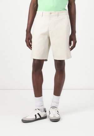 Beige cotton shorts with a straight cut, featuring a front button closure, side pockets, and a smooth texture. Worn with white sneakers.