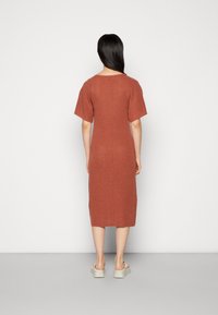 Frau mit langen dunklen Haaren, die ein rostfarbenes, knielanges Strickkleid und weiße Plateausandalen trägt, vor einem schlichten weißen Hintergrund stehend.