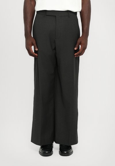 Pantalons larges gris foncé sur mesure en tissu lisse, avec une taille plate, des passants de ceinture et des poches latérales. Associés à des chaussures noires.