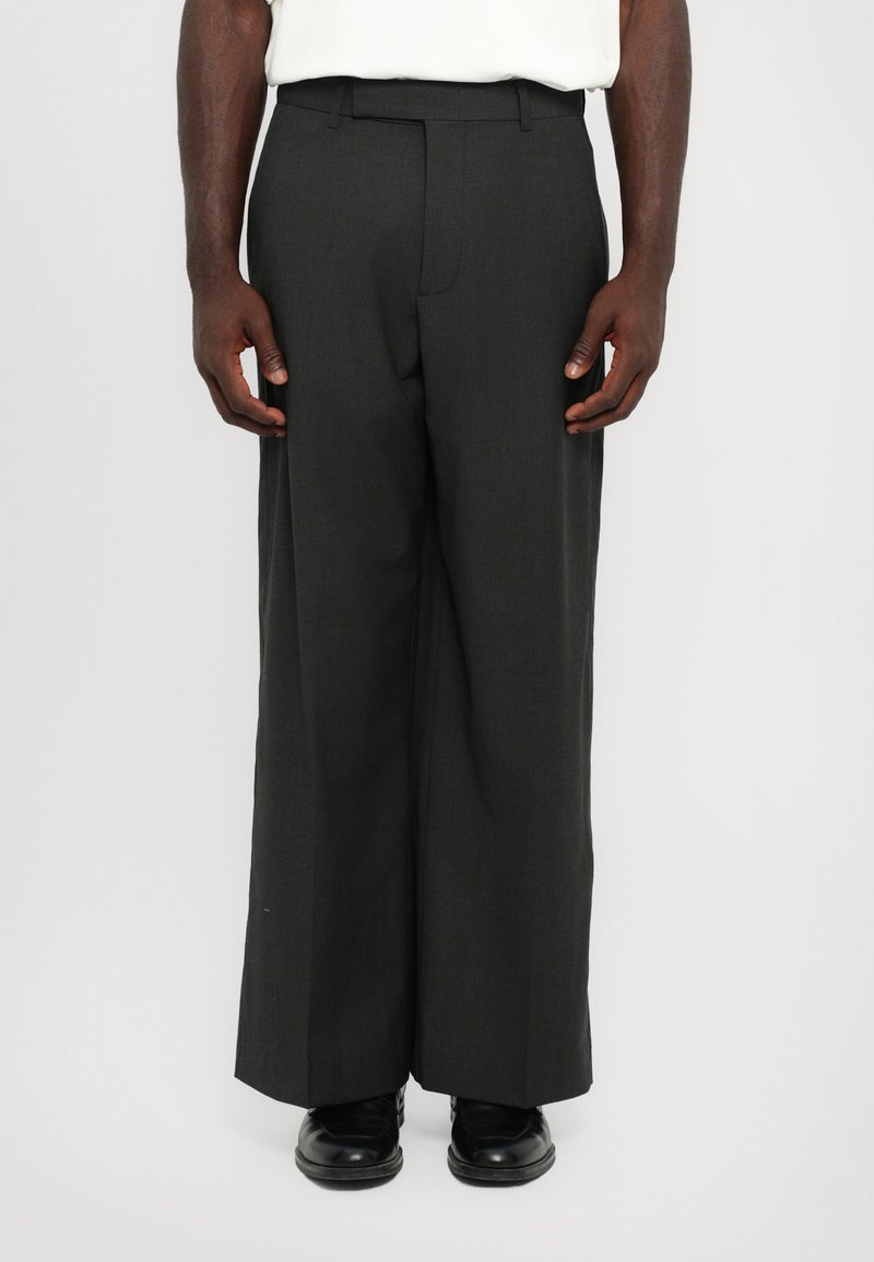 Pantaloni a gamba larga grigio scuro su misura, realizzati in tessuto morbido, con vita piatta, passanti per cintura e tasche laterali. Abbinati a scarpe nere.