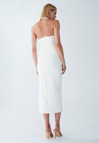 Weißes trägerloses Midikleid mit einer taillierten Oberkörperpartie und einem Reißverschluss auf der Rückseite, kombiniert mit hautfarbenen High-Heels. Glatte Stofftextur.