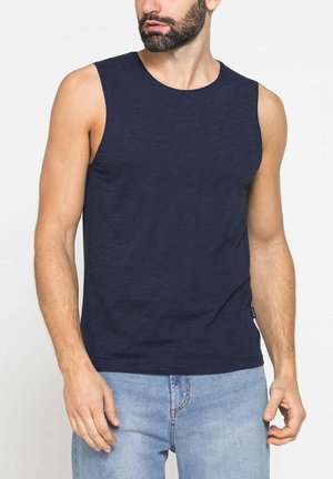 Uomo con barba che indossa una canotta blu navy e jeans azzurri, in piedi davanti a uno sfondo bianco semplice.