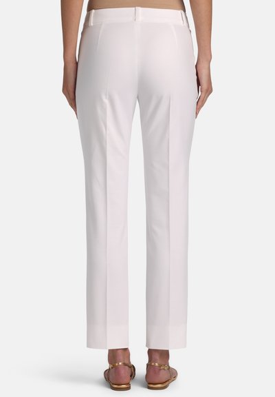 Back view of a person wearing straight-leg white trousers and gold open-toe sandals standing against a plain background.